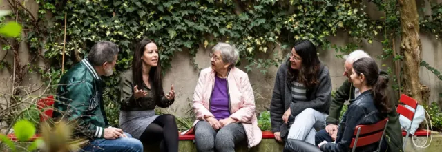 Eine Gruppe von sechs Personen unterschiedlichen Alters sitzt im Kreis in einem grünen Garten und hört aufmerksam einer Frau zu, die mit ausdrucksstarken Gesten spricht.