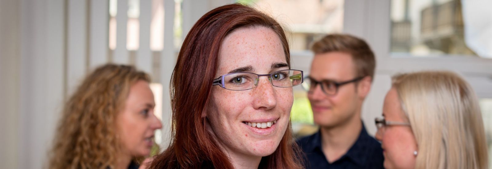 Eine Frau mit Brille blickt in die Kamera, ein Mann und zwei Frauen unterhalten sich im Hintergrund.